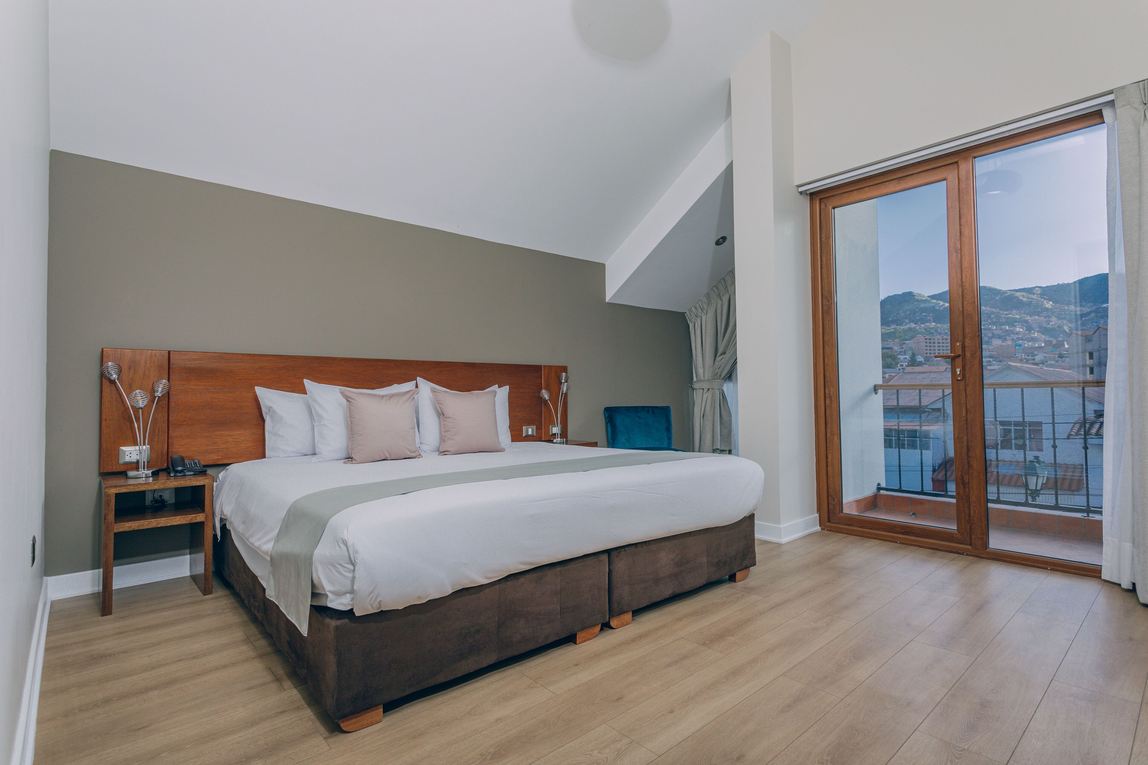 Un dormitorio con una cama grande, una mesita de noche, un cabecero de madera y un ventanal corredero que da a un balcón con vistas a la ciudad.
