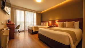 Ein Hotelzimmer mit zwei Betten, einem Schreibtisch, einem Sessel, einem Fernseher und einem großen Fenster mit Vorhängen.