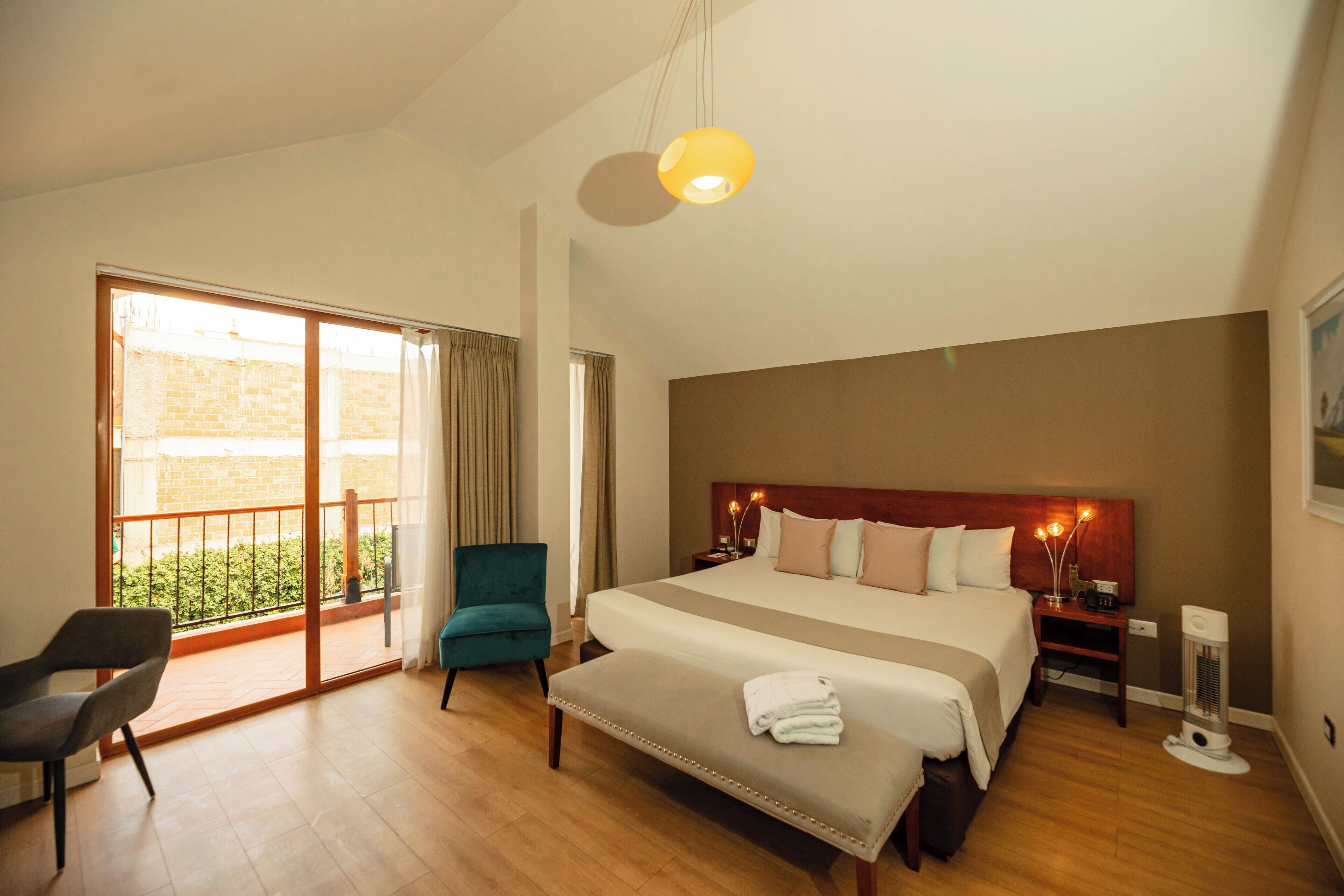 Un dormitorio espacioso con una cama grande, un banco, una silla, una mesita de noche con lámparas y vistas a un área exterior por una puerta corredera.