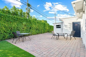 Une terrasse avec une table et des chaises, une haie et un bâtiment avec une porte.
