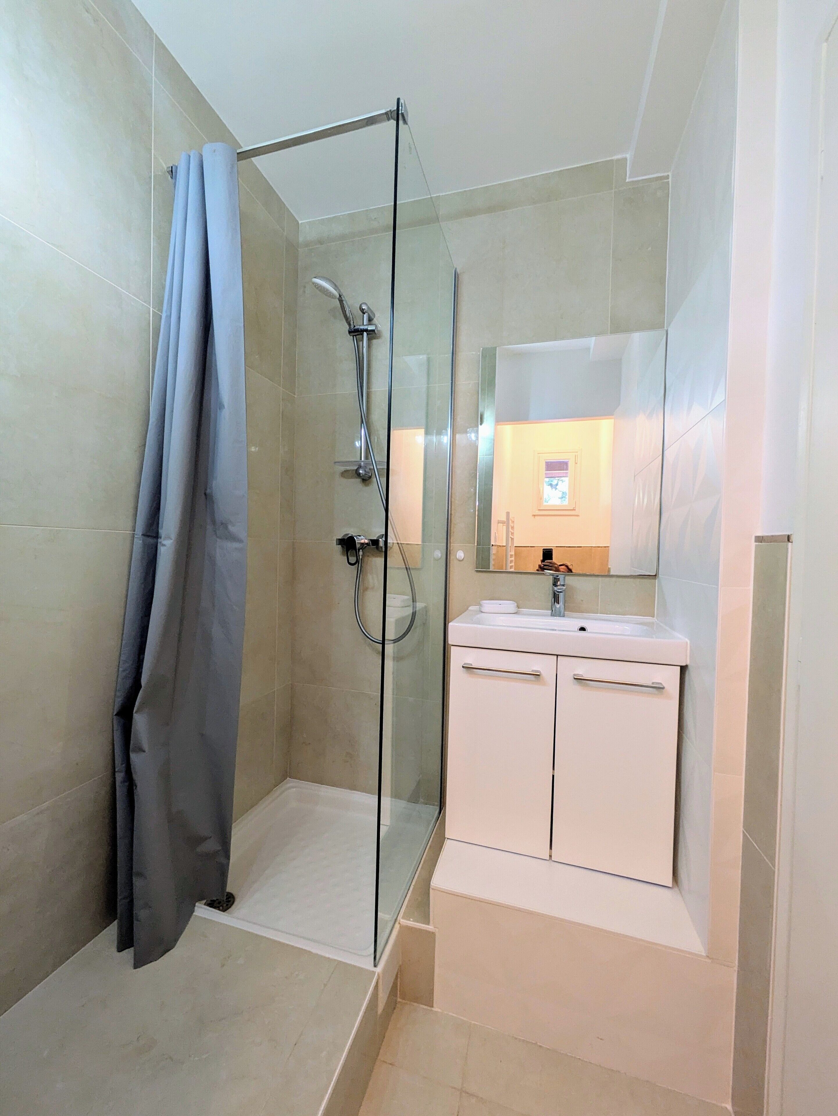 Une salle de bain moderne avec une douche à parois de verre, un meuble-lavabo blanc avec un miroir et des murs recouverts de carreaux de couleur beige.