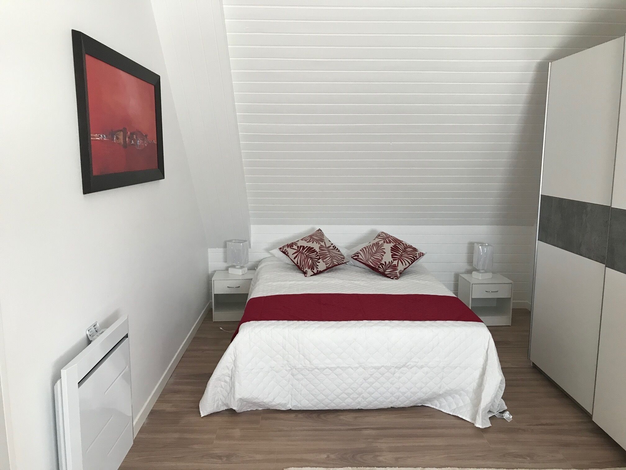 Een slaapkamer met een bed, twee nachtkastjes, een radiator, een kledingkast en een abstract schilderij in rood op de muur.