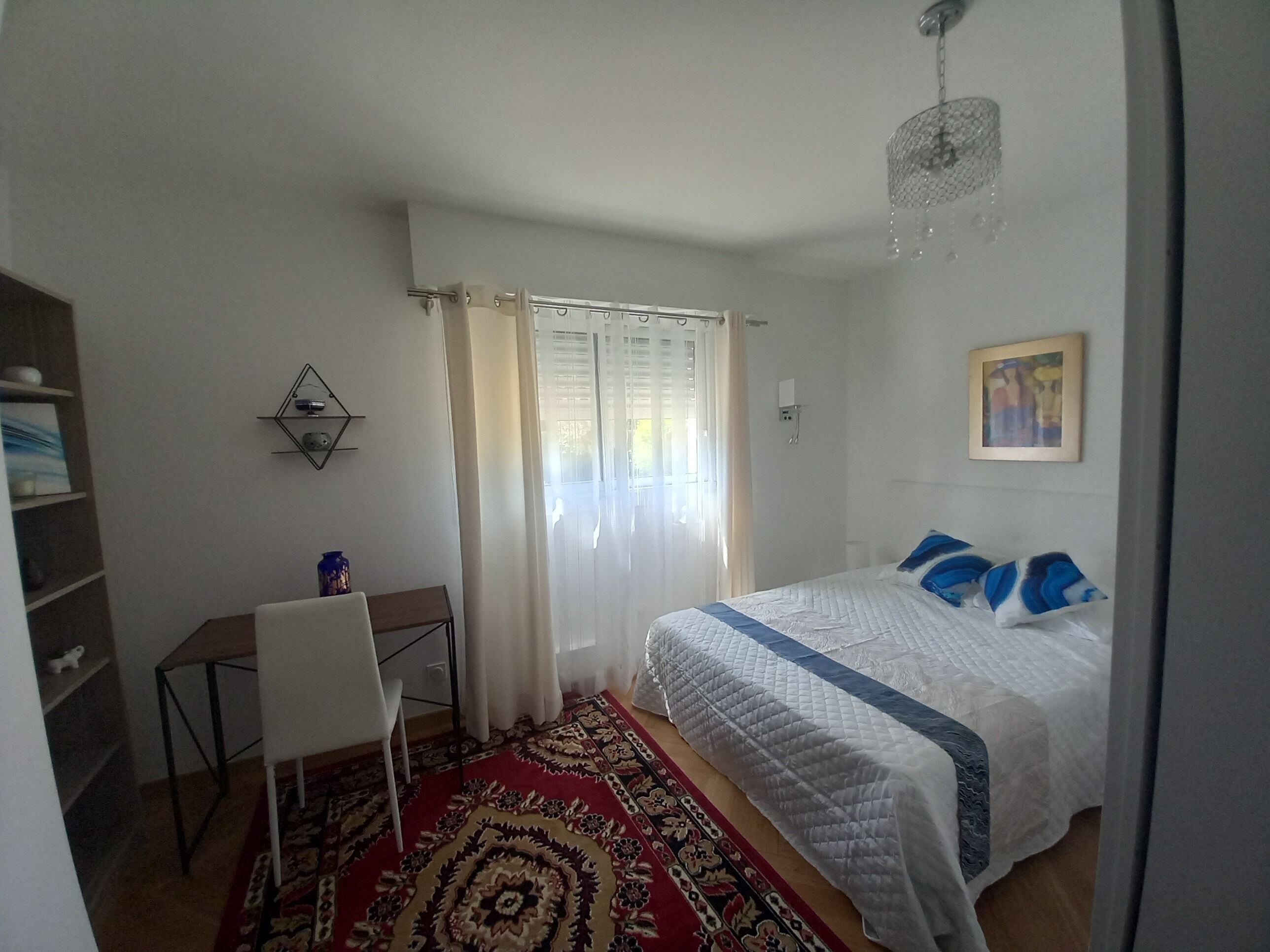 A bedroom with a bed, a desk, a chair, a rug, a chandelier, and a painting.