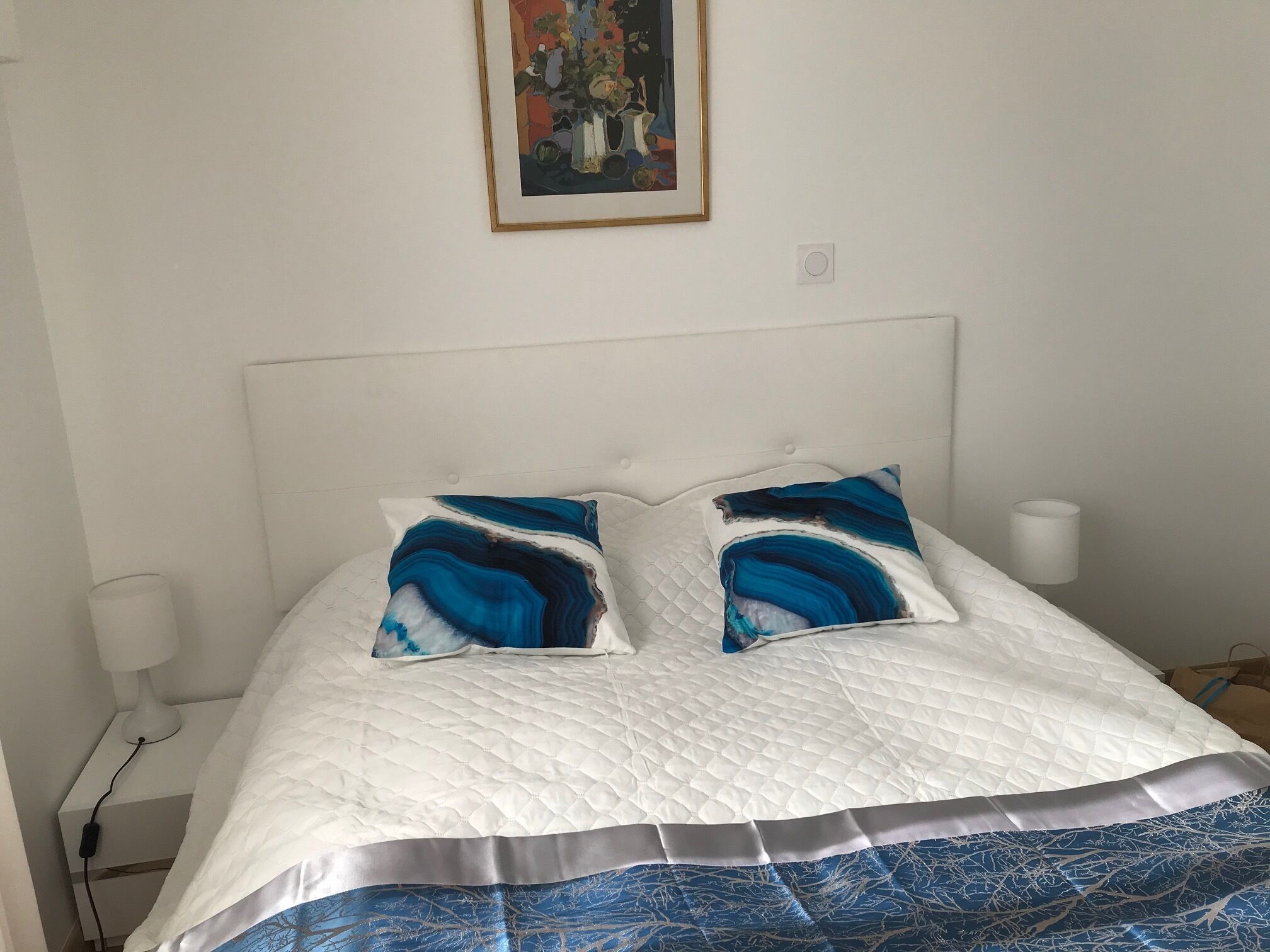 Un lit avec une tête de lit blanche matelassée, deux coussins décoratifs, une table de chevet blanche avec une lampe, et un tableau accroché au mur.
