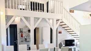 A spacious living area with a high ceiling, a staircase, and a dining area with white furniture.