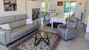 A modern living room with a sofa, armchair, and coffee table. A dining area with a table and chairs is visible in the background.