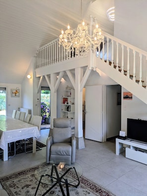 A spacious living room with a high ceiling, a chandelier, a dining area with a table and chairs, a grey armchair, and a staircase leading to an upper level.