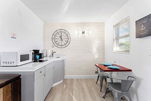 Une cuisine compacte équipée d’un four à micro-ondes, d’une machine à café et d’un évier. Une horloge est fixée au mur, et une fenêtre avec des stores est visible.
