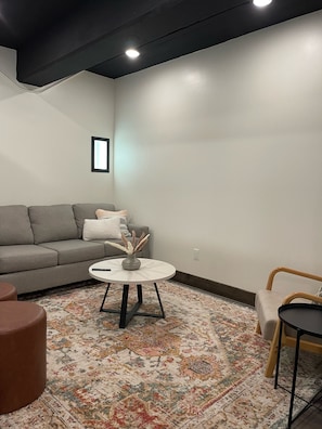 Ein modernes Wohnzimmer mit einem grauen Sofa, einem runden Couchtisch mit einer Vase, einem gemusterten Teppich und einem Holzstuhl.