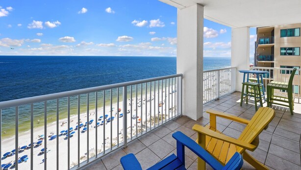 A balcony with a view of the beach and ocean, featuring a railing, chairs, and tables.