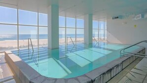 An indoor swimming pool with clear glass walls, large windows, and a view of the beach.