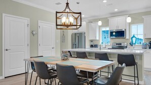 A modern kitchen with white cabinets, a large island, and a dining area with chairs and a table. - New Townhome with Private Beach Club (Pensacola)
