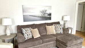 A living room with a sectional sofa, two side tables, and a large landscape painting on the wall. - Tranquil Home in Beautiful Setting (Ames)