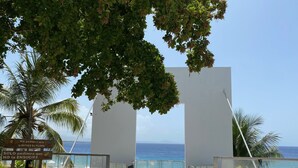 Entrada de un resort con vista despejada al océano, palmeras y un letrero con un mensaje.