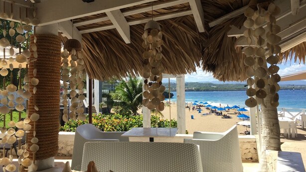 Un espacio de relax frente a la playa con mobiliario de mimbre, techo de paja y vistas al mar.