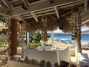 Eine Strandlounge mit Korbmöbeln, einem Reetdach und Blick auf Strand und Meer.