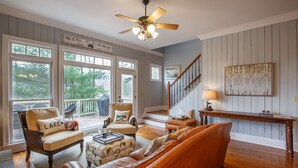 A spacious living room with a large window, a ceiling fan, a wooden staircase, and a view of the outdoors. - Lake's Edge Lodge - Water Views, & Community Golf! (Big Canoe)