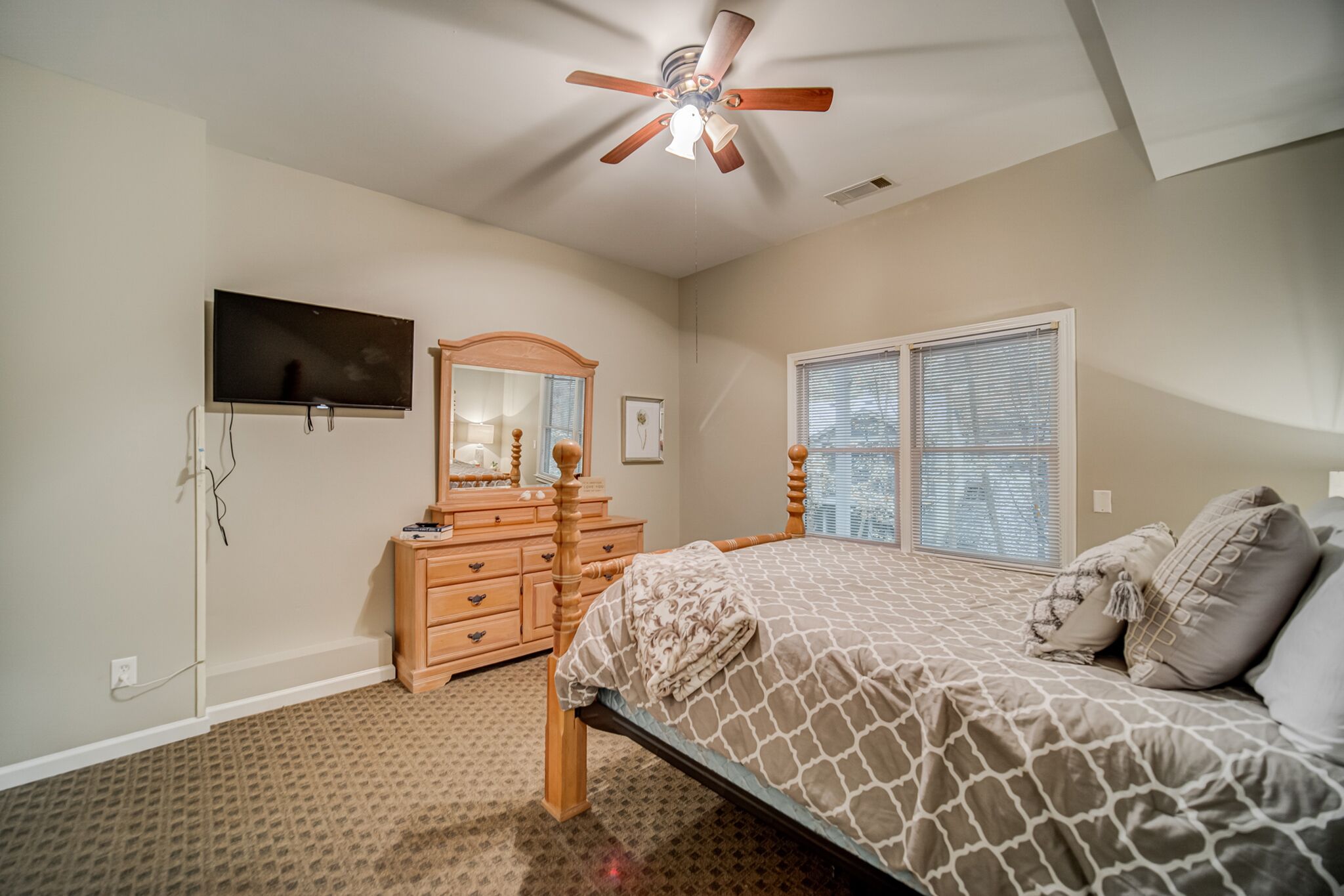 A bedroom with a bed, dresser, mirror, ceiling fan, and a TV mounted on the wall.