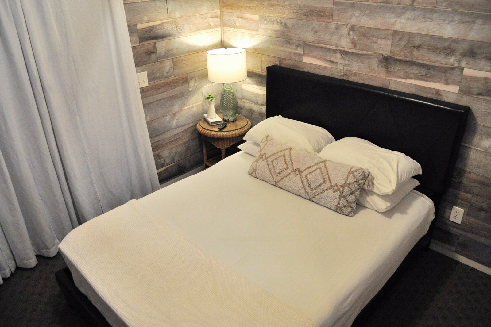 Una cama bien hecha con un cojín estampado, una lámpara de noche y un panel de pared de madera.