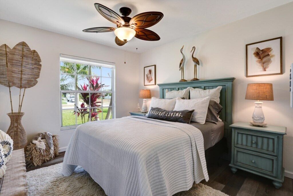 A bedroom with a bed, a ceiling fan, a nightstand, a lamp, and a window with a view of plants outside.