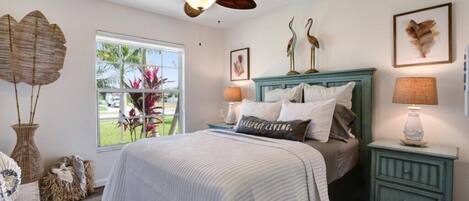 A bedroom with a bed, a ceiling fan, a nightstand, a lamp, and a window with a view of plants outside.