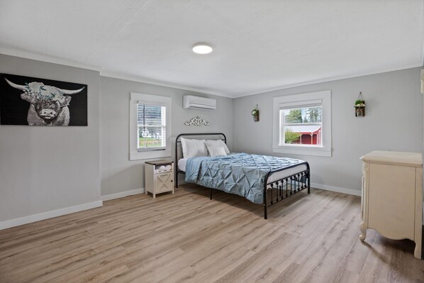 A bedroom with a bed, bedside tables, a wall-mounted air conditioner, and two windows with blinds.