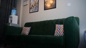 A green sofa with patterned cushions, a water dispenser, and two framed artworks on the wall.