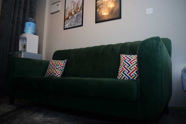 A green sofa with patterned cushions, a water dispenser, and two framed artworks on the wall.