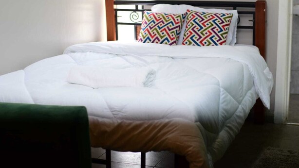 A neatly made bed with white bedding and colorful patterned pillows, a wooden headboard, and a green chair in the foreground.