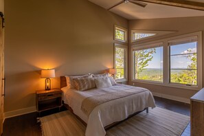 Un dormitorio con una cama grande, dos lámparas de noche, una mesita de noche y vistas al exterior a través de amplias ventanas.