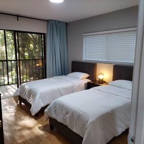 Un dormitorio con dos camas, una ventana con persianas, una cortina y vistas al exterior con vegetación.