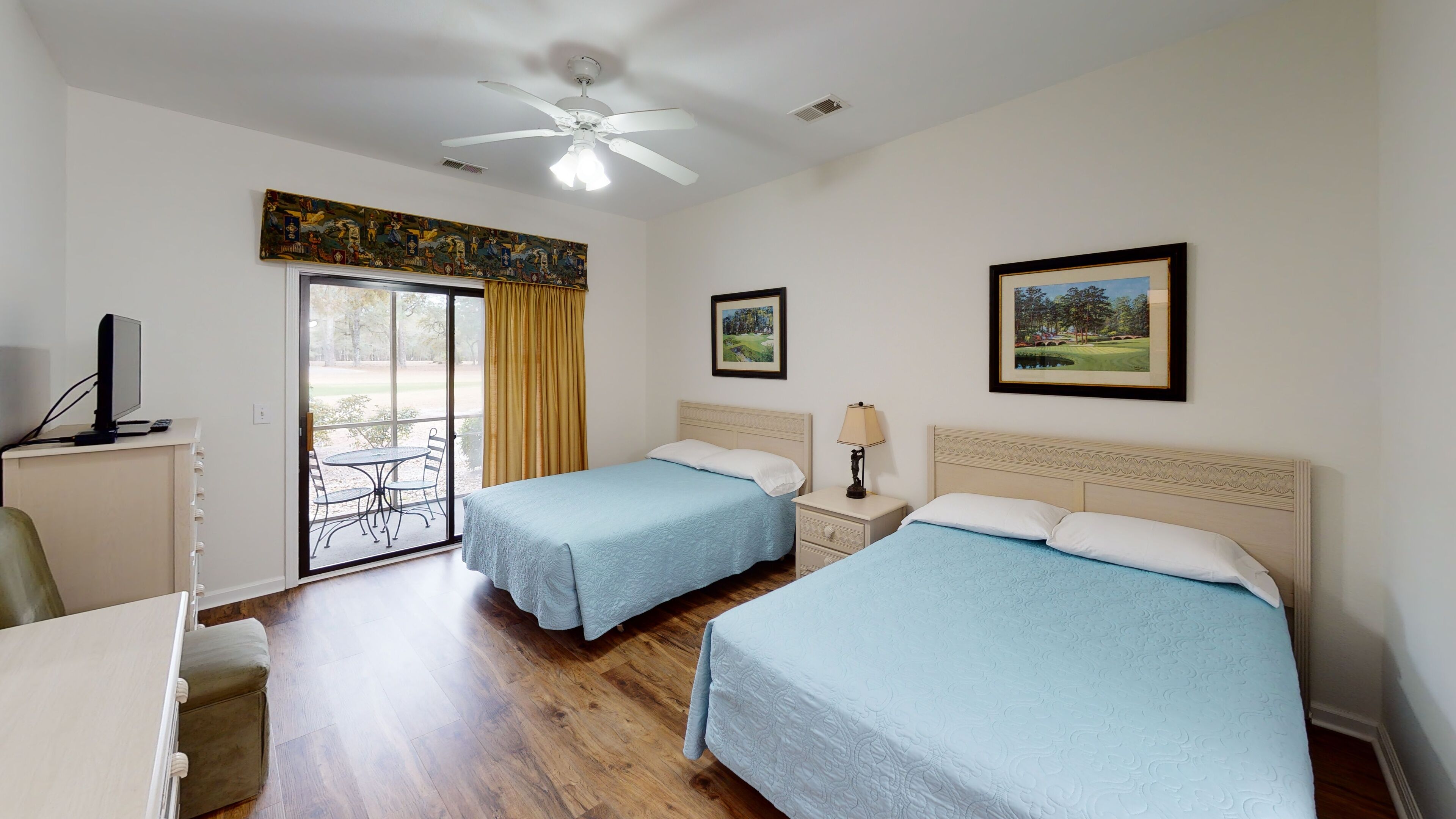 A hotel room with two beds, a desk, a chair, a TV, and a balcony with a view of trees.