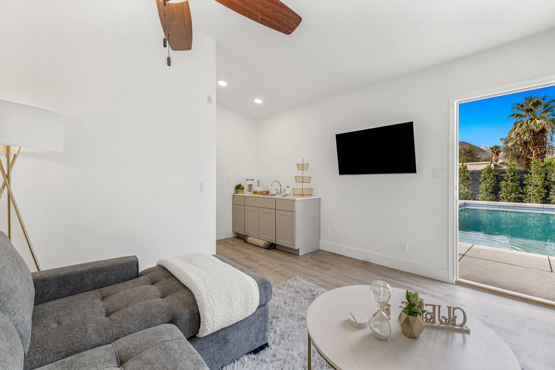 A modern living room with a grey sofa, a round coffee table, and a kitchen area with a sink and cabinets. A flat-screen TV is mounted on the wall, and a pool is visible through the glass door.