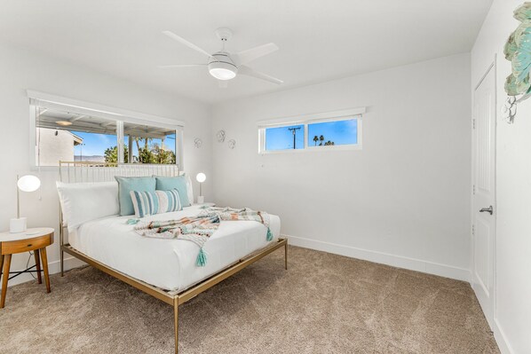 A bedroom with a bed, a ceiling fan, a window with a view, and a nightstand with a lamp. - Heated Pool & Spa & Games, Family & Pet Friendly, (Palm Desert)