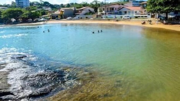 A beach with clear water, a sandy shore, and buildings in the background. - Pousada Boa Ventura (Anchieta)