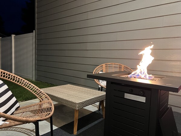 An outdoor patio with a table and two wicker chairs, a built-in fire feature, and a wooden fence in the background.