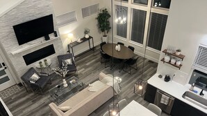 Un salon moderne avec un canapé, une table basse et un téléviseur. Il y a également un coin repas avec une table et des chaises. La cuisine est équipée d’un évier et de placards.