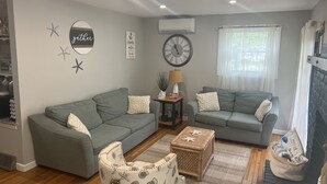 Un salon chaleureux avec deux canapés gris, une petite table avec une lampe, une horloge murale et un mur décoratif orné d’étoiles de mer et d’ancres.