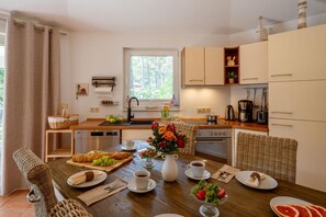 A modern kitchen with a wooden table set for breakfast, a sink, and a window overlooking greenery.