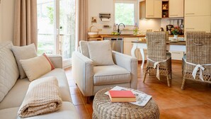 Una sala de estar moderna con un sofá, sillones y un otoman de mimbre. Cuenta con una zona de cocina con armarios y una mesa de comedor.
