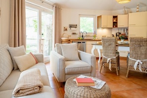 A modern living room with a sofa, armchairs, and a wicker ottoman. There is a kitchen area with cabinets and a dining table.