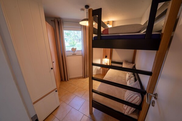 A compact room with a bunk bed, a small window with curtains, a radiator, and a door.