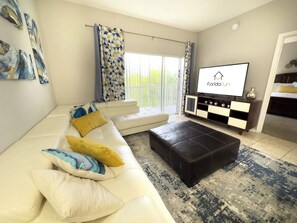 A modern living room with a white sofa, a black ottoman, a TV on the wall, and a balcony with patterned curtains.