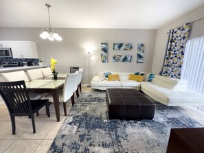 A modern living room with a white sofa, a dining table with chairs, and a kitchen in the background.