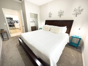 A bedroom with a large bed, a wooden headboard, and a nightstand with a lamp. There is a closet with a hanging rack and a small table with a plant.