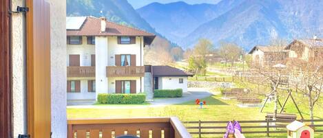 A balcony with a table and chairs, overlooking a mountain landscape with a playground and a small house.