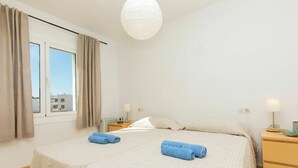 Une chambre moderne avec un grand lit, deux tables de chevet, un luminaire suspendu et une vue sur les bâtiments par la fenêtre.