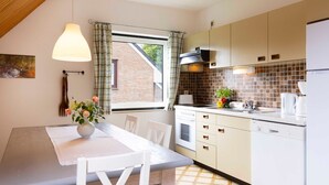 A modern kitchen with a dining area, featuring a white table, chairs, a vase with flowers, a window with curtains, and a brick wall visible through the window. - (STOE3) Holiday apartment "Waack" - Holiday apartment "Waack" up to 4 persons (Timmendorfer Strand)