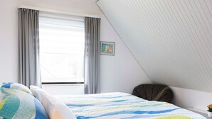 A bedroom with a bed, two pillows, a window with curtains, a painting on the wall, and a slanted ceiling. - (STOE3) Holiday apartment "Waack" - Holiday apartment "Waack" up to 4 persons (Timmendorfer Strand)