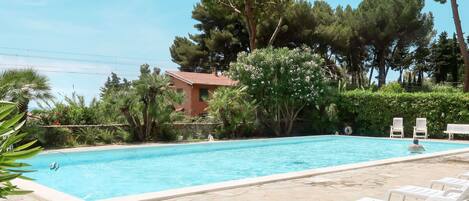 A swimming pool with stone tiles, surrounded by white lounge chairs, and a house with a red roof in the background.